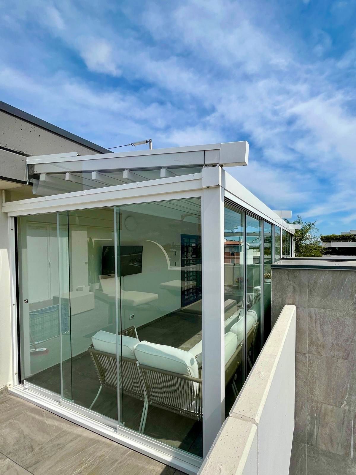 Terrazza panoramica in vetro con struttura bianca e sedute imbottite sotto un cielo azzurro.