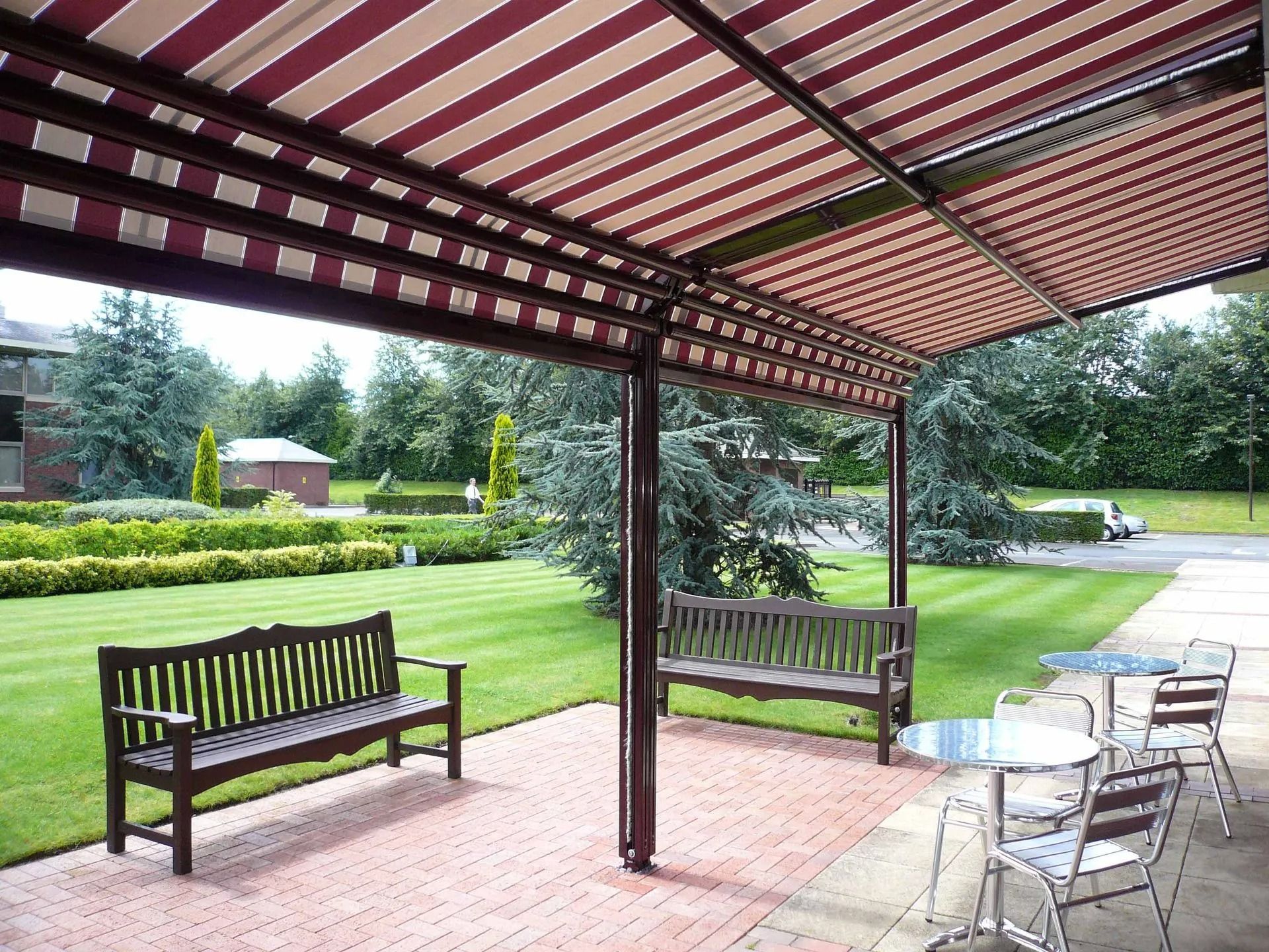 Patio coperto con tetto a strisce rosse, panchine e un tavolino che si affaccia su un giardino curato.