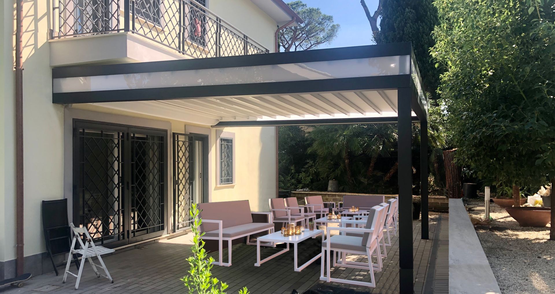 Patio posteriore con tavolo da pranzo all'aperto sotto un pergolato nero accanto a una casa color crema.
