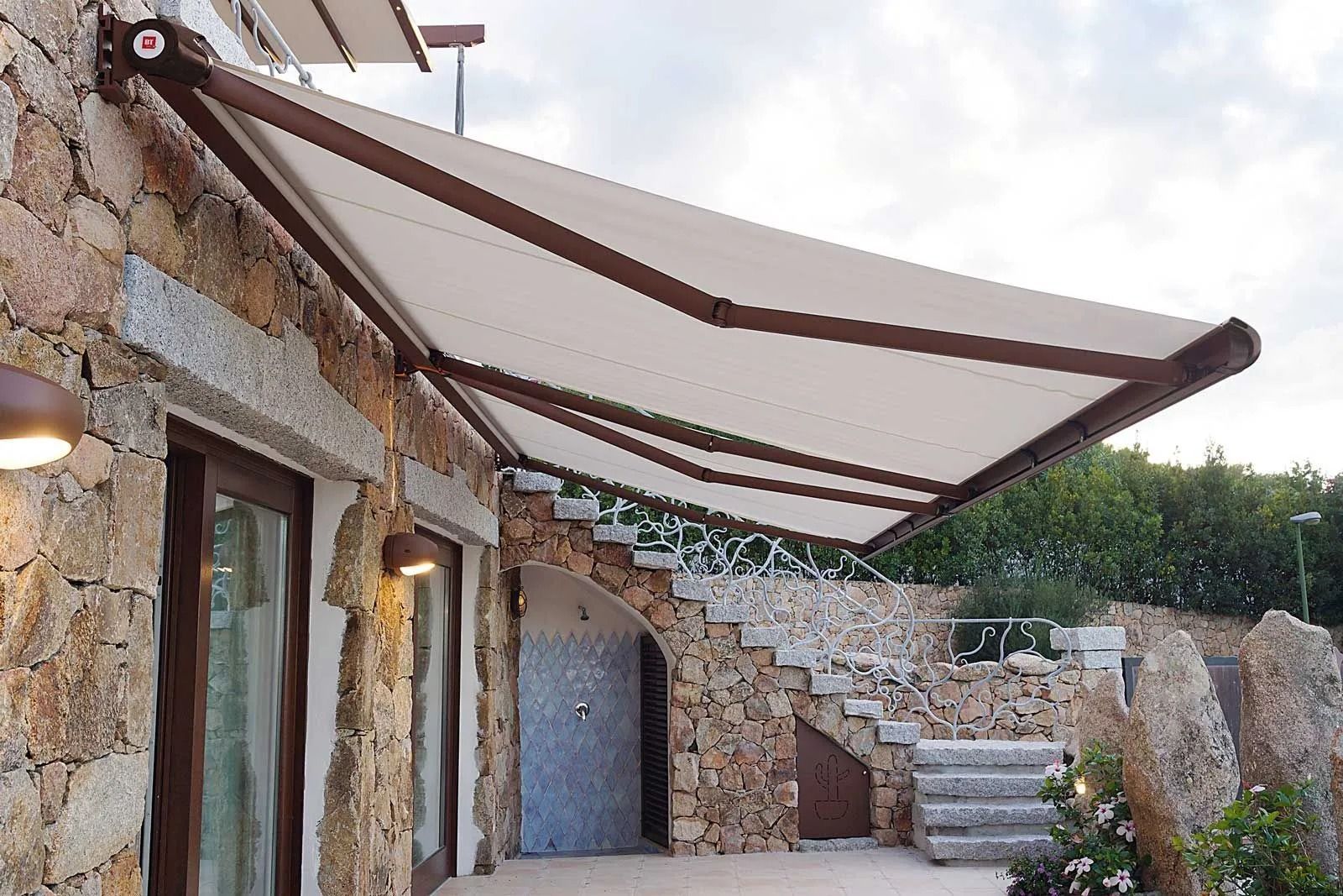 Terrazza in pietra con un'ampia tenda da sole beige sopra edifici rustici e scalini sotto un cielo nuvoloso