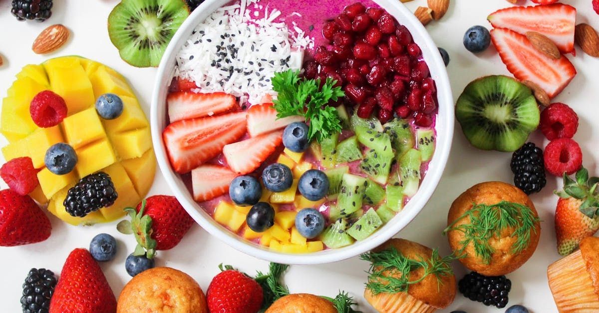 Un bol de fruta está rodeado de otras frutas y verduras sobre una mesa.