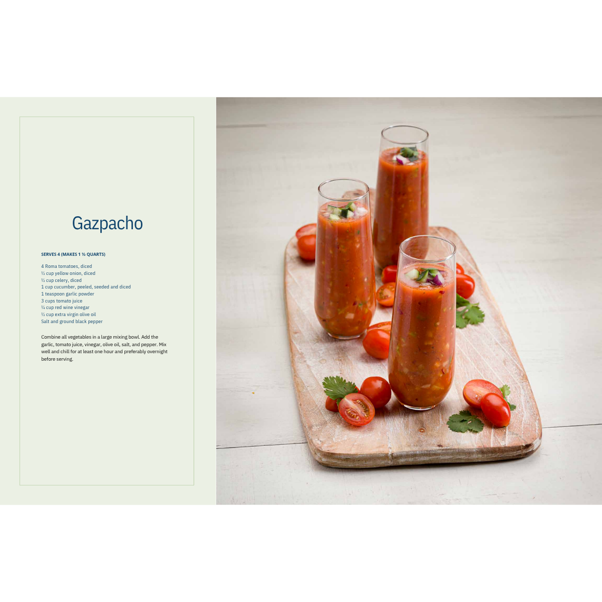 Gazpacho served in tall glasses on a tray with strawberries and herbs, beside a recipe page.
