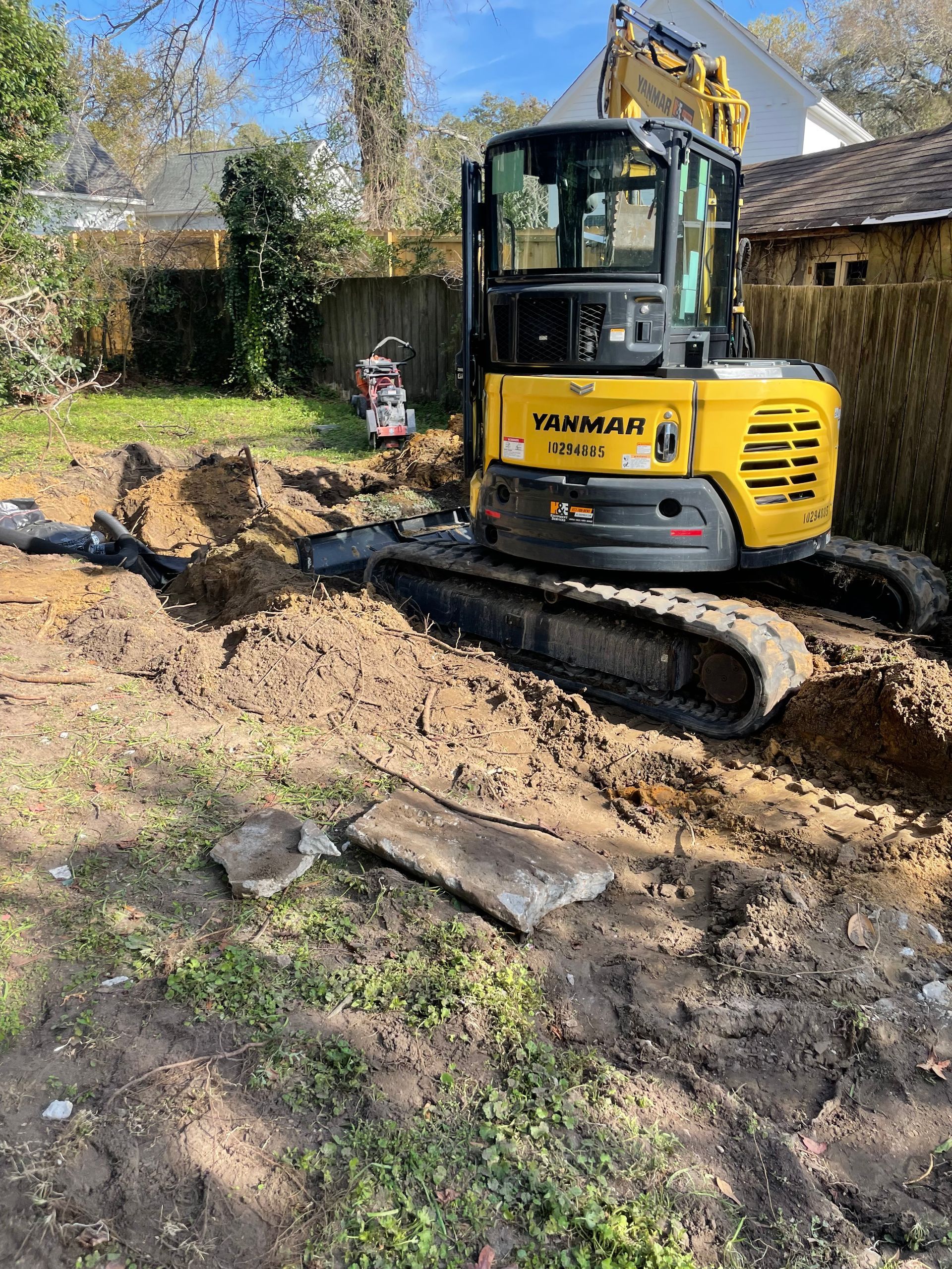 A yellow excavator is digging a hole in the dirt in a backyard.