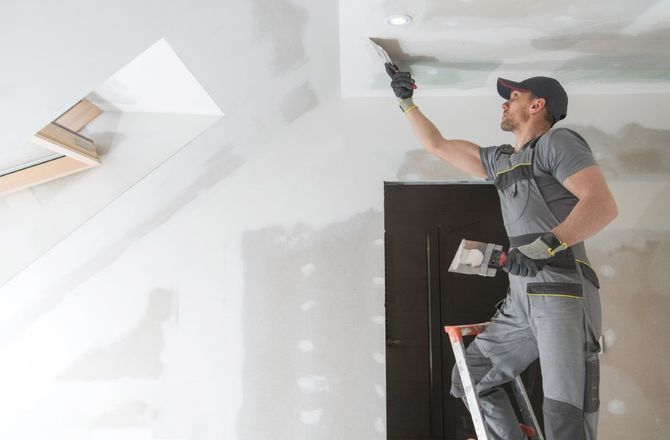 Worker painting a white ceiling with a roller while standing on a ladder.