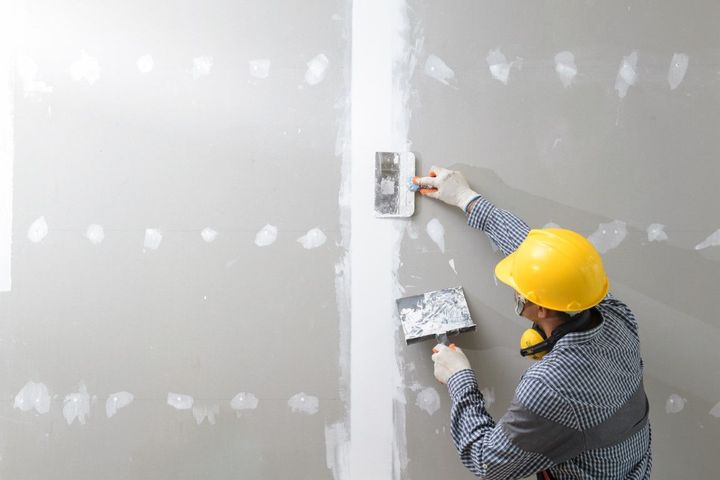 Worker in yellow hard hat plastering and smoothing a gray wall with a trowel and float.