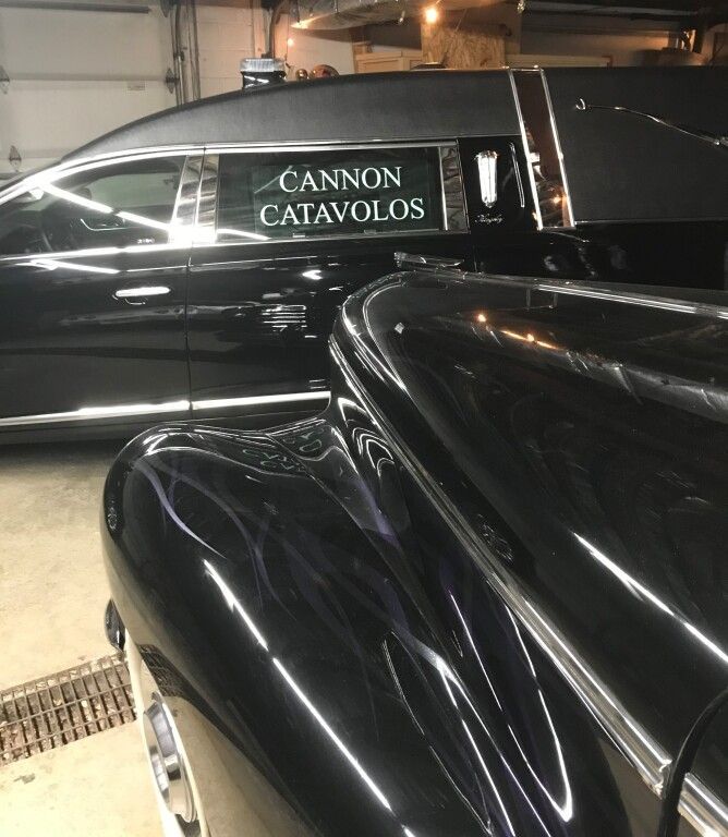 Black hearse parked near a classic black car inside a garage. The hearse has a