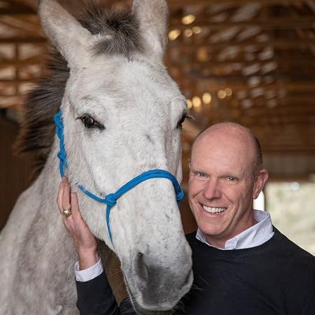 Smiling man in black sweater standing beside a white donkey with a blue halter in a barn