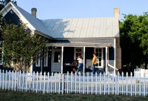 A blue, single-story house with a white picket fence, a metal roof, and people standing on the front porch.