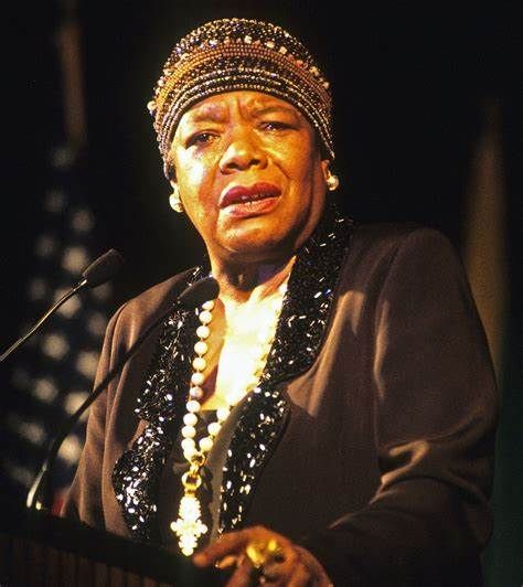 Woman at a podium, wearing a beaded head wrap and necklace, speaking into a microphone.