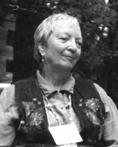 Woman with short hair, wearing a vest with floral embroidery, smiling outdoors. White badge hanging from neck.