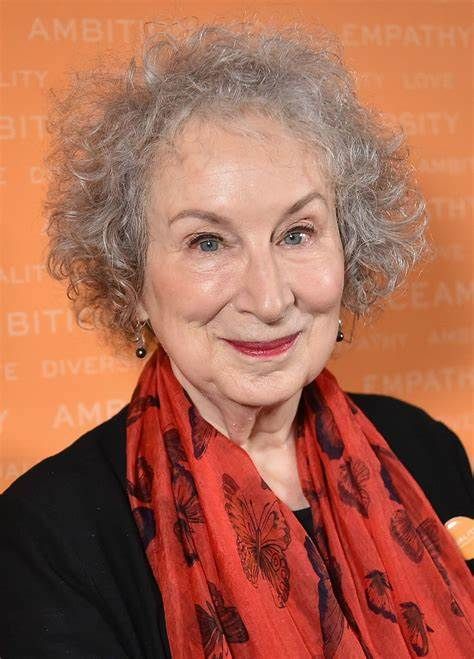 Margaret Atwood smiles, wearing a red scarf, black top, and has curly grey hair.