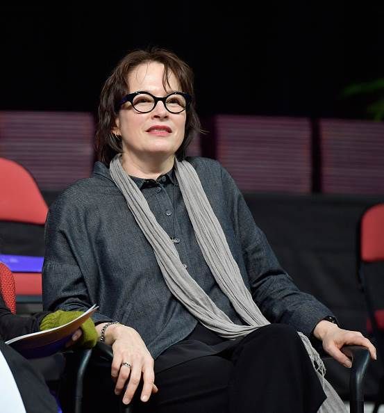 Woman with glasses, sitting, wearing a gray scarf and jacket. Background is blurred.