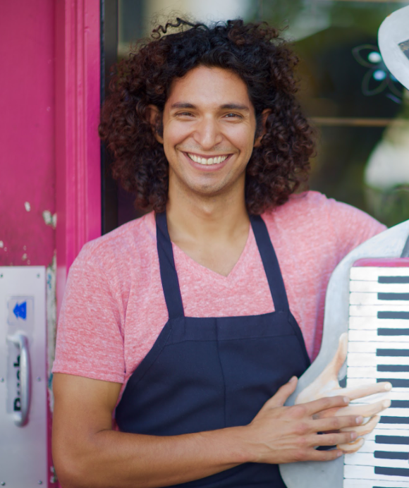 Man with curly hair, smiling, wearing an apron, holding an accordion, standing in a pink doorway.