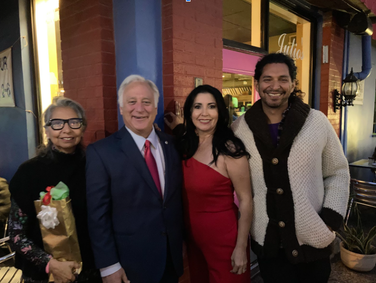 Four people pose outdoors: a woman holding flowers, a man in a suit, a woman in a red dress, and a man in a sweater.