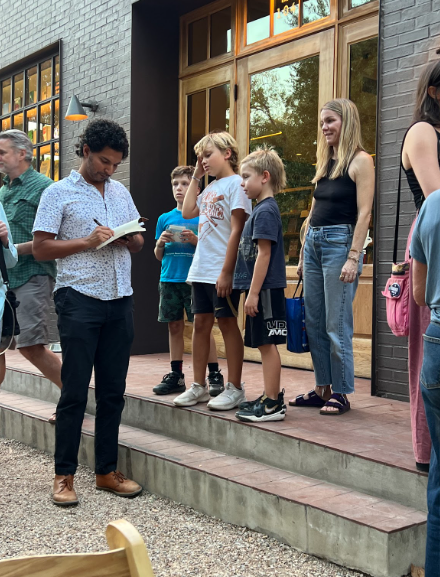 Person signing an autograph for children outside a building. Other people observe nearby.