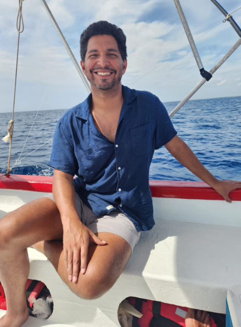 Man on a sailboat, smiling, wearing a blue shirt and shorts, sitting on the boat's edge with ocean background.