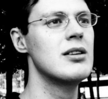 Man with glasses looking upward, slight smile, outdoor setting.