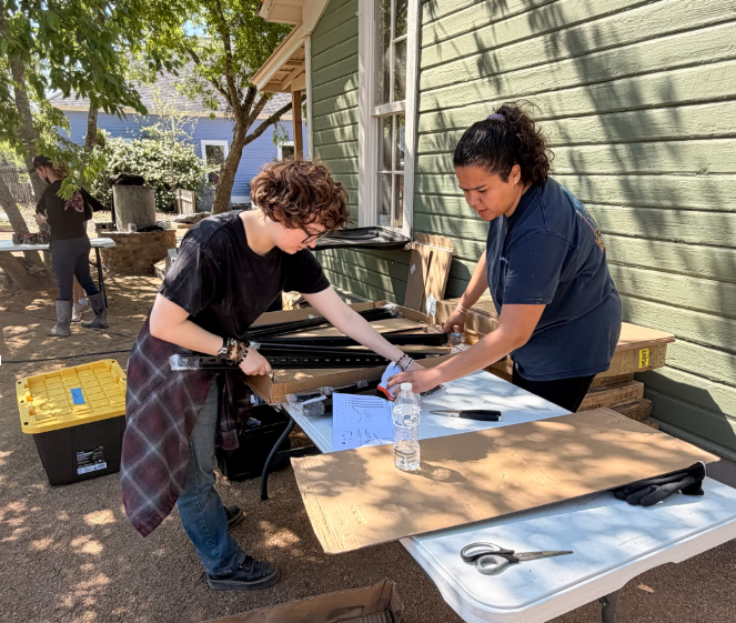 Two people sorting materials at an outdoor worktable beside a house