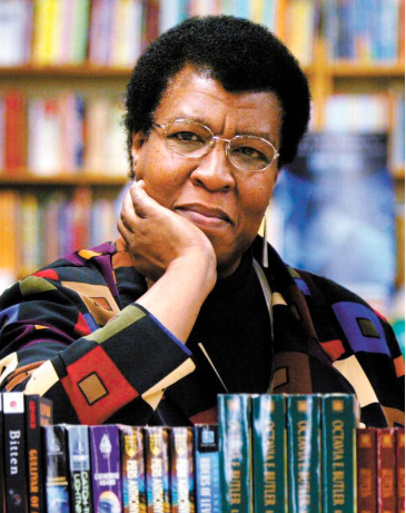 Woman with glasses resting chin on hand, in front of a bookshelf with books by Octavia Butler.