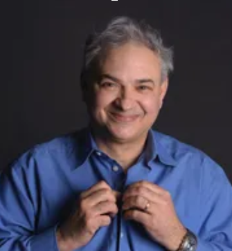Man in a blue shirt buttoning it up, smiling against a dark background.