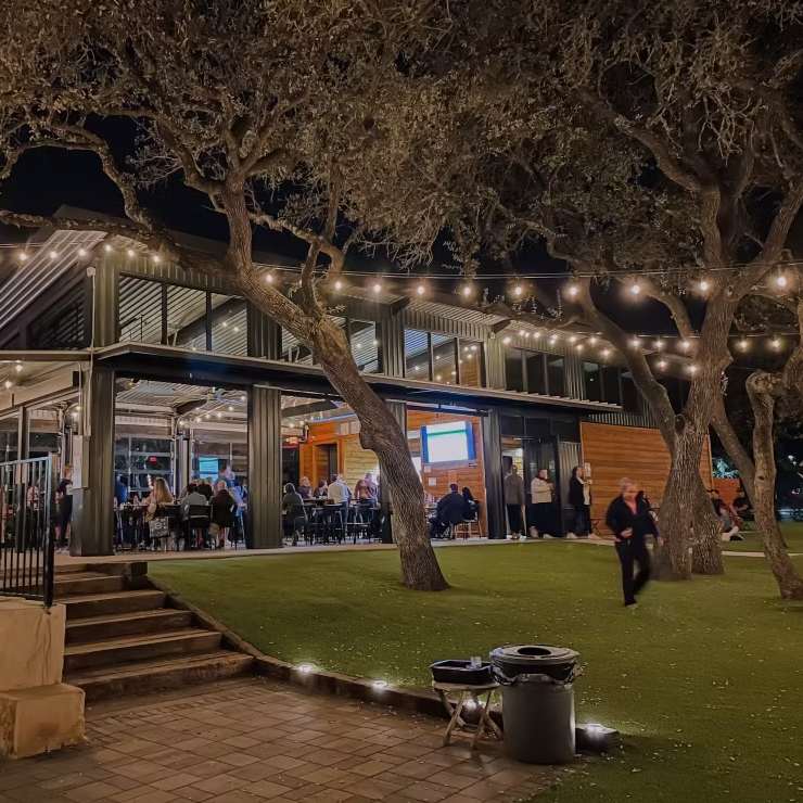 Nighttime outdoor restaurant patio with string lights, trees, and seated diners behind glass walls.