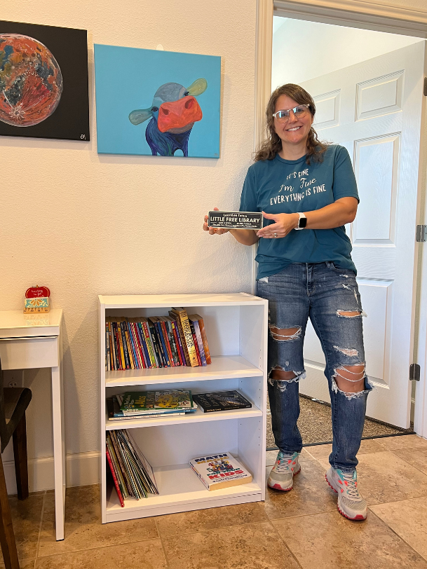 Woman holding object by white bookshelf with books, art on wall.