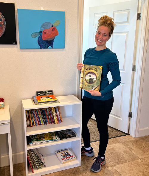 Woman holding a book, smiling near a bookshelf with children's books. Artwork of a cow on the wall.