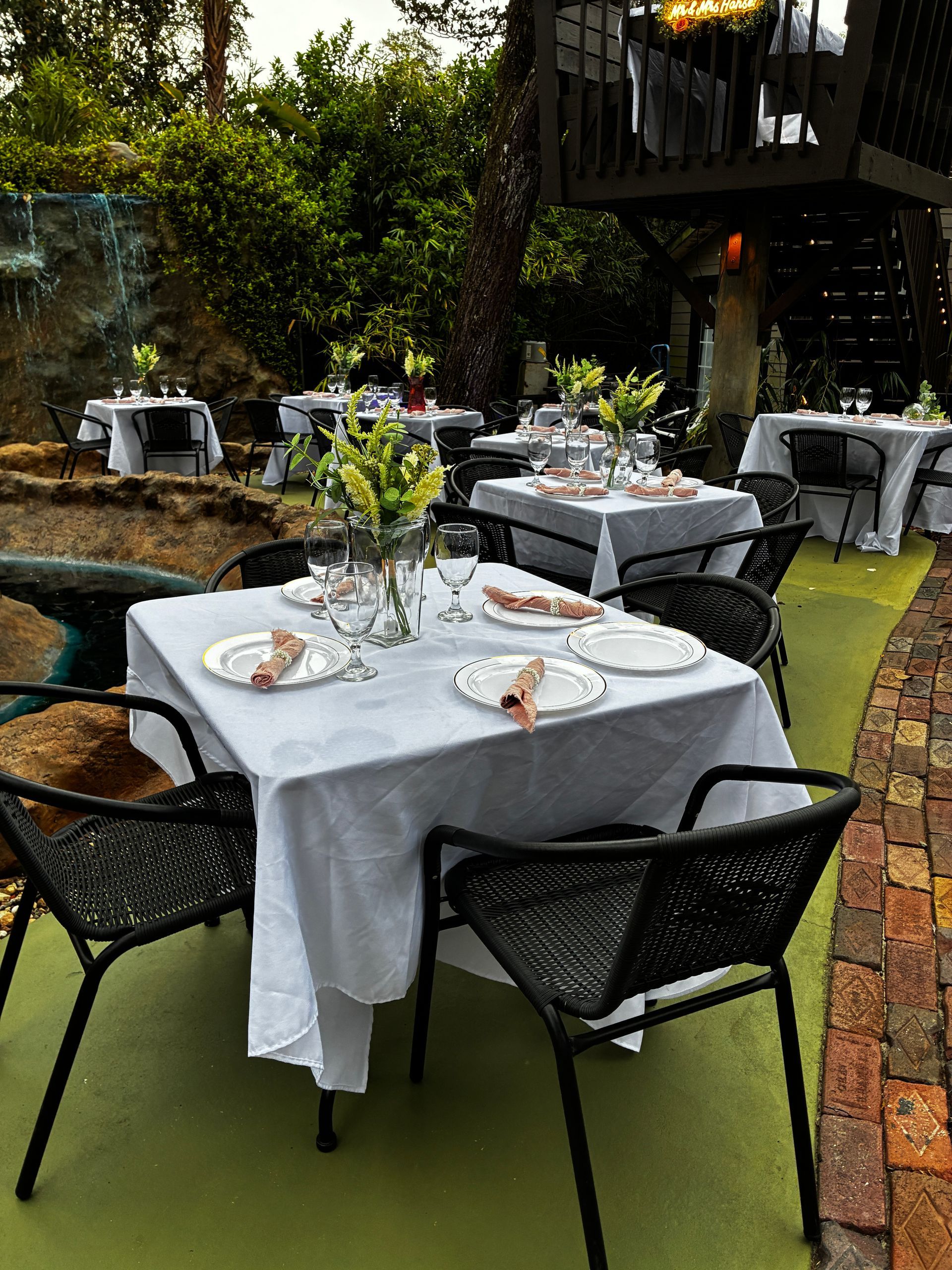 Outdoor restaurant tables with white tablecloths beside a pond and green lawn chairs, set for dining