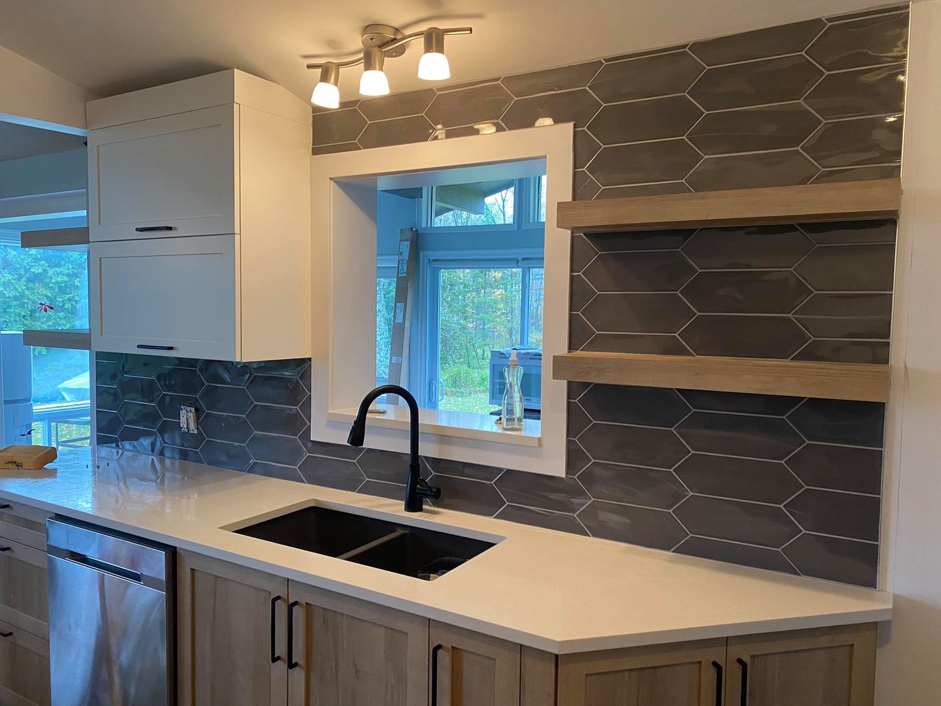 A kitchen sink with a matte black faucet, dark gray picket tile backsplash, white countertop, and wooden floating shelves.