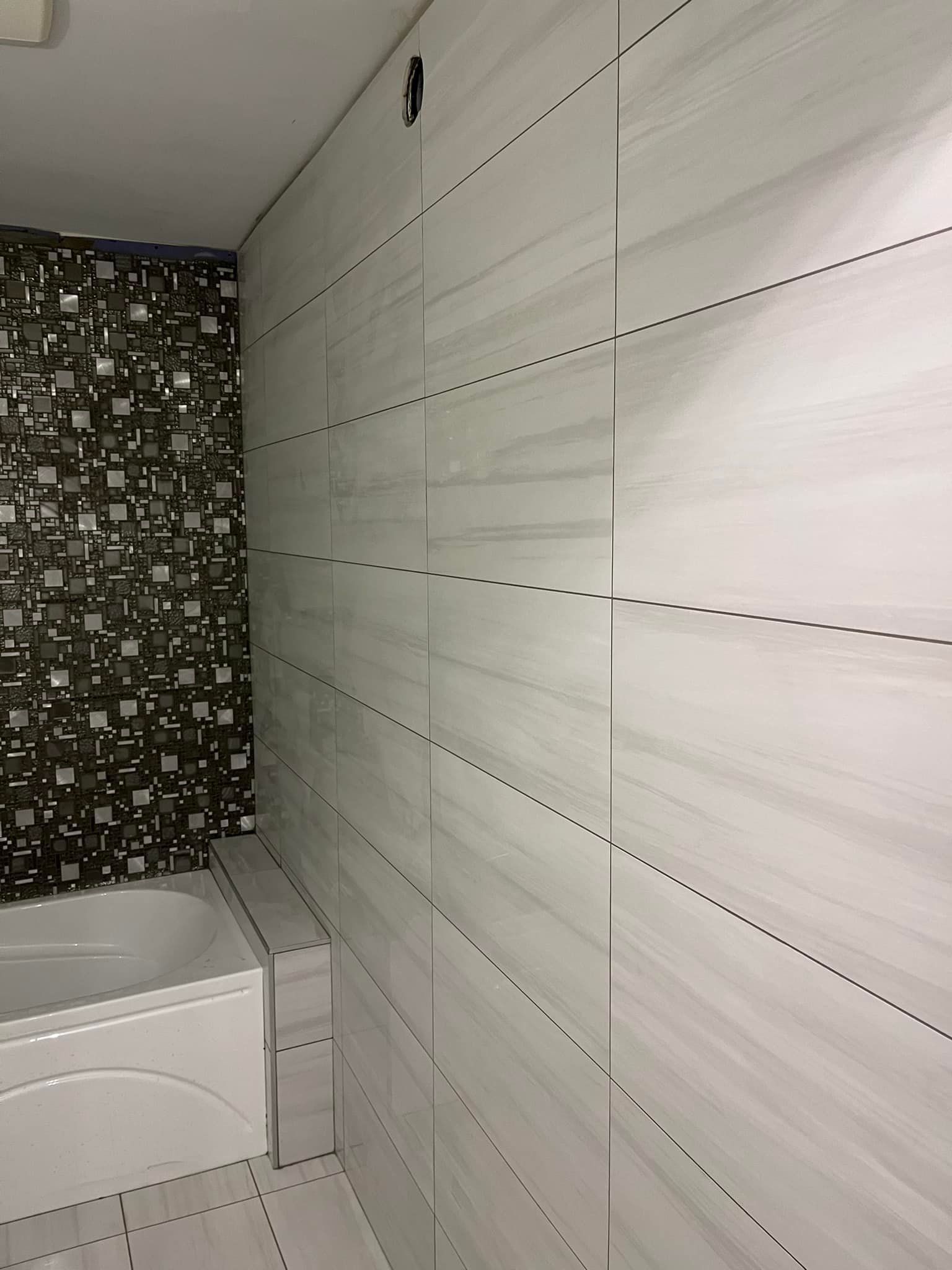 Une salle de bains avec une baignoire blanche adossée à un mur recouvert de carreaux horizontaux gris clair et d'une section de mosaïque décorative.