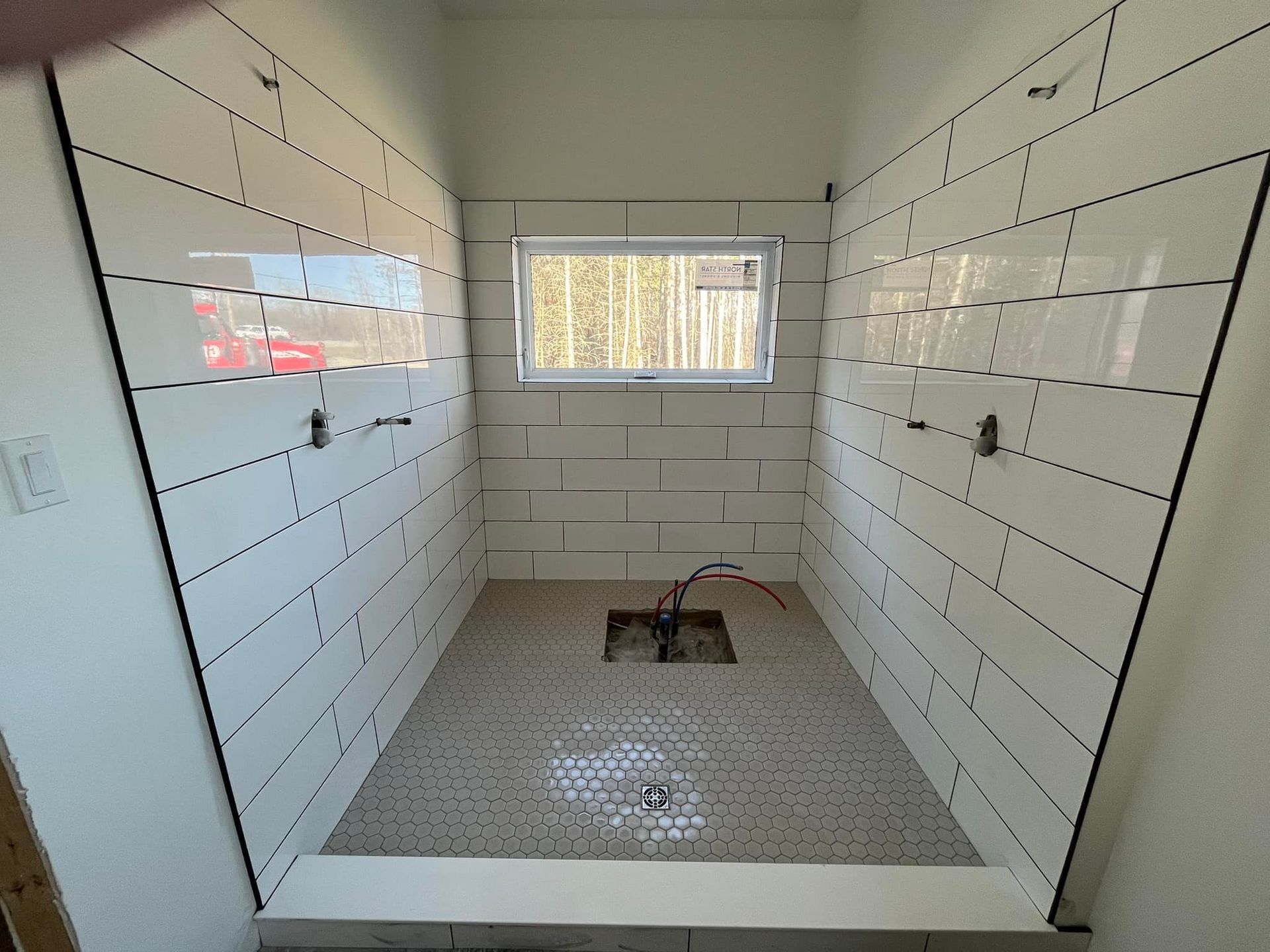 Une douche à l'italienne en construction avec des carreaux de métro blancs aux murs, une petite fenêtre et un sol en mosaïque grise.