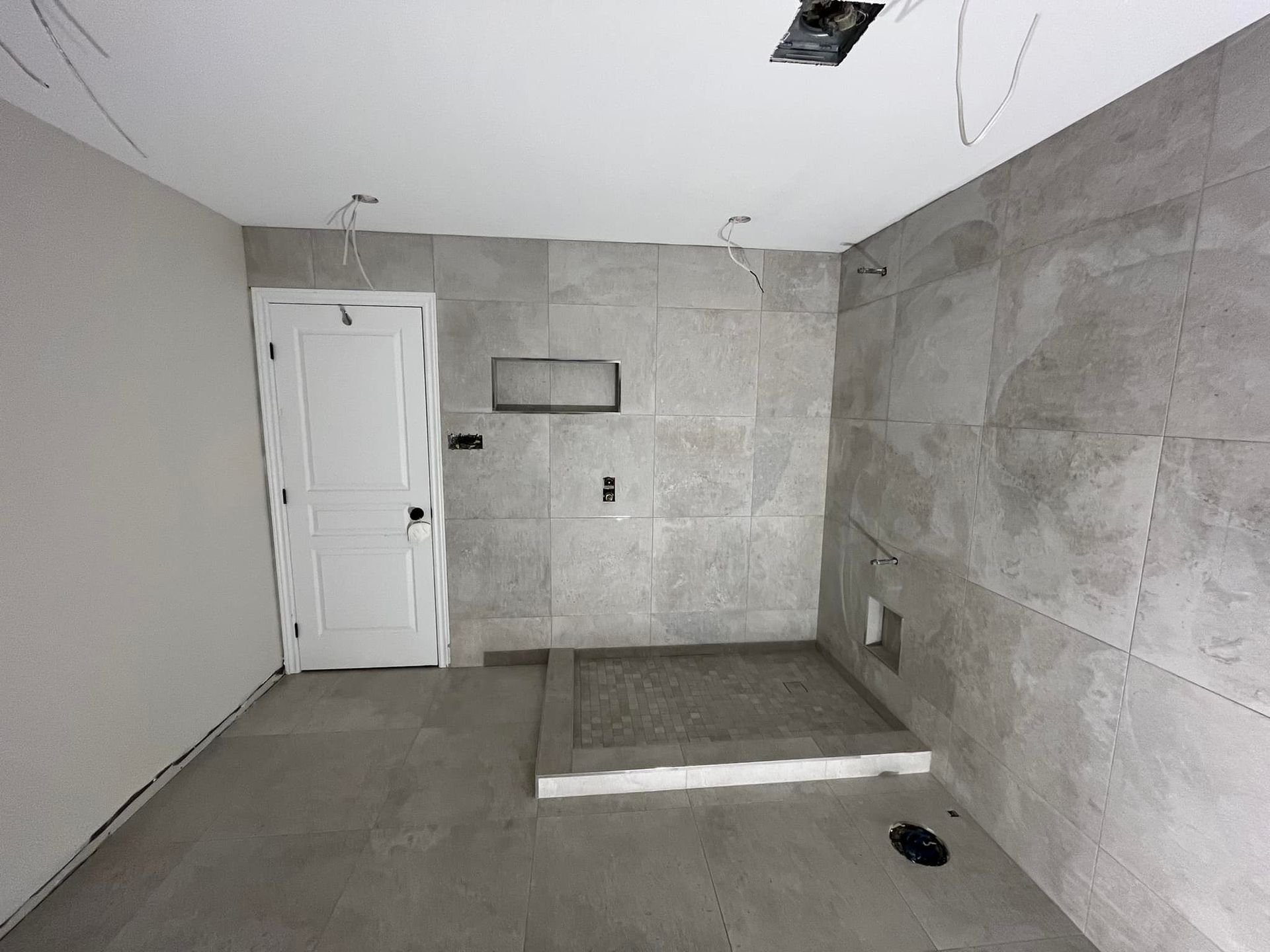 Une salle de bains en construction avec des carreaux de pierre grise aux murs et au sol, une porte blanche et un receveur de douche surélevé.