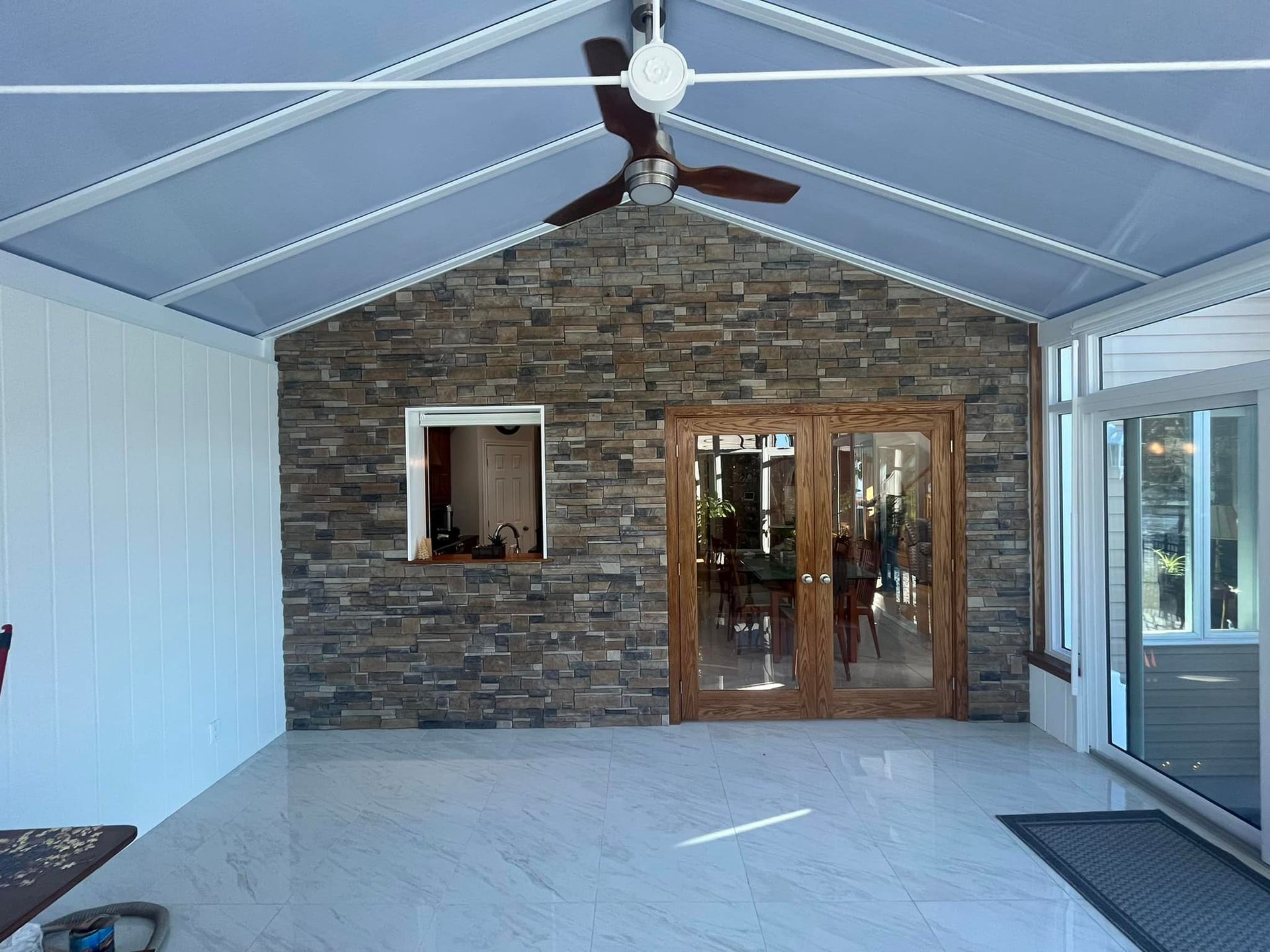 Un porche en pierre avec un ventilateur de plafond, des lambris blancs, un sol carrelé blanc et des portes doubles vitrées donnant sur l'intérieur.