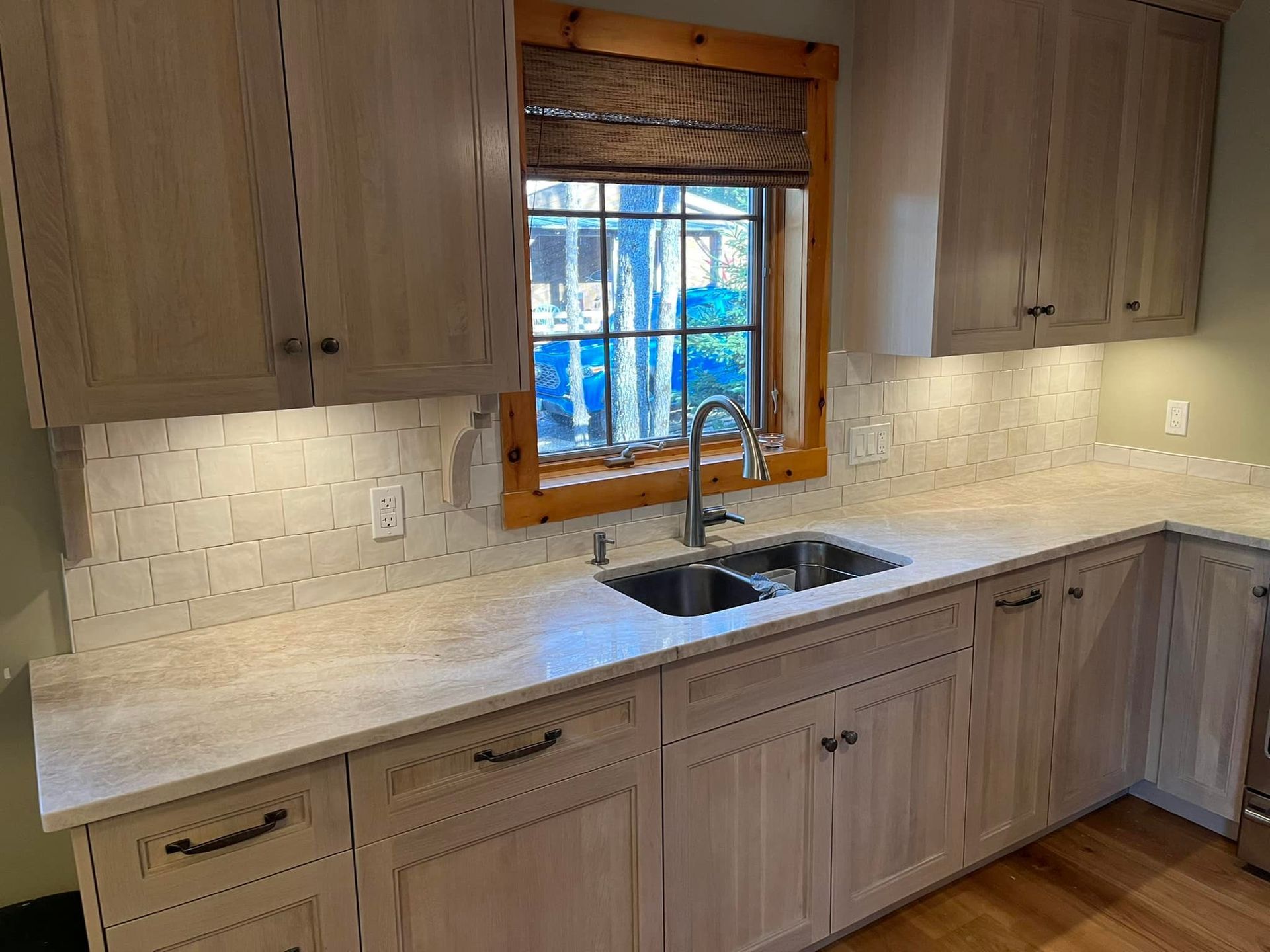 Cuisine claire avec armoires en bois, comptoirs en quartz, crédence en carrelage et évier sous une fenêtre à cadre en bois.