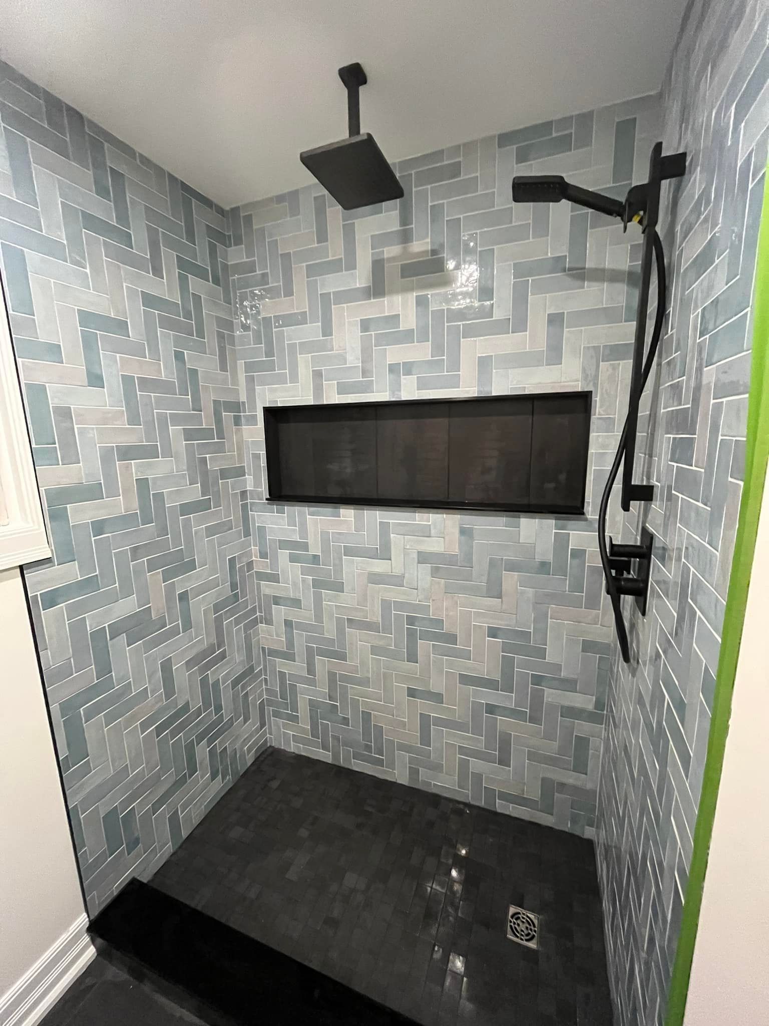 Une douche avec des murs carrelés de carreaux à motifs bleus et blancs, une pomme de douche noire, une niche noire et un sol en pierre sombre.