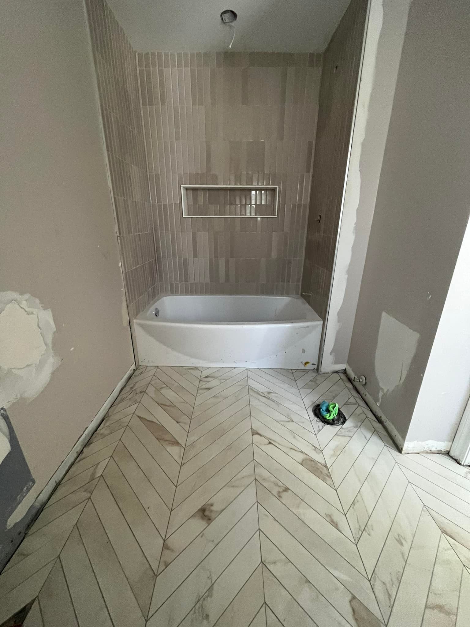 Une salle de bains en cours de rénovation comprenant une baignoire blanche, un sol carrelé à chevrons dans des tons clairs et un mur de douche carrelé beige avec une niche.
