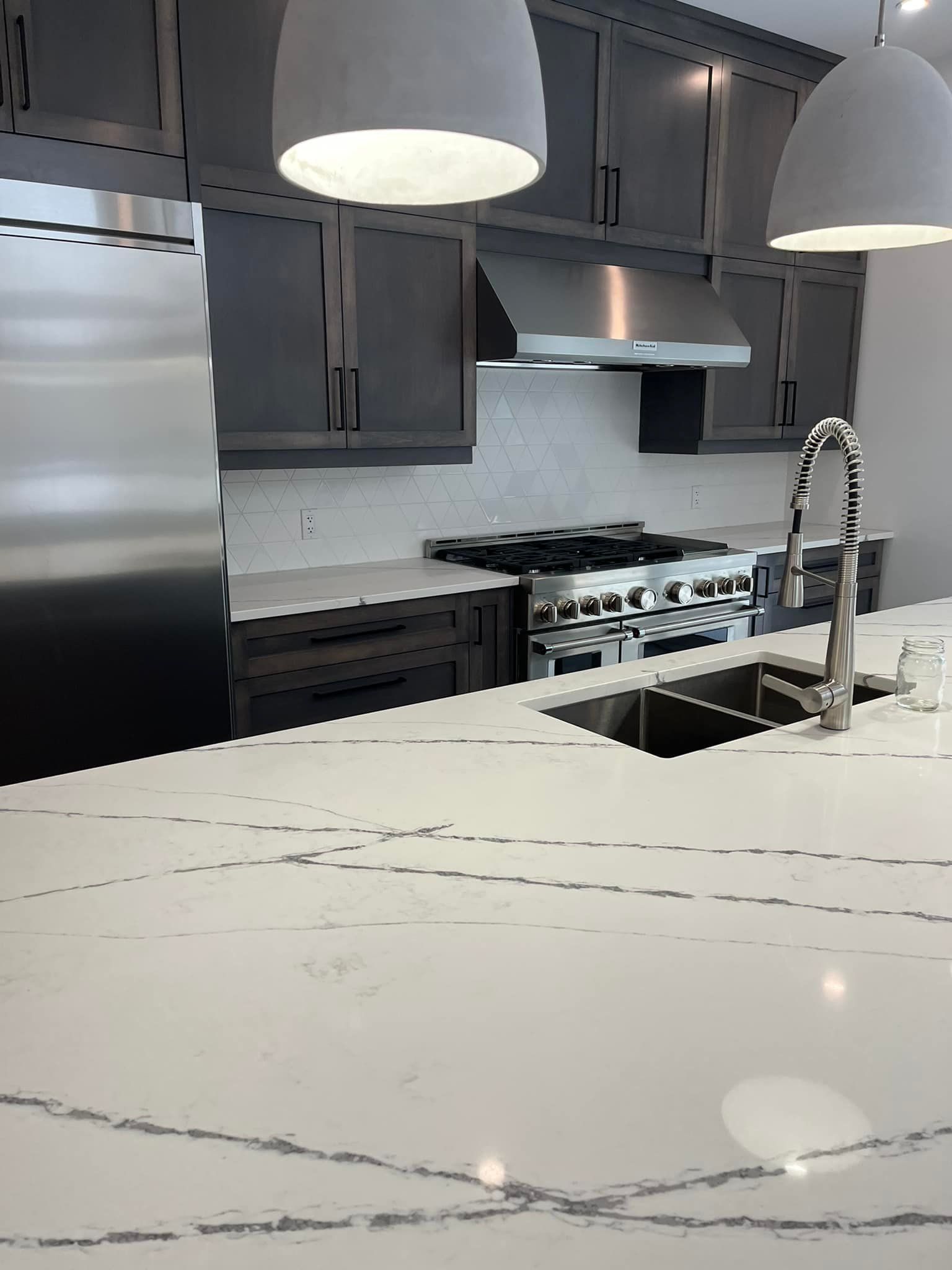 Cuisine moderne avec un îlot central en marbre blanc à motifs, des armoires gris foncé, des appareils électroménagers en acier inoxydable et des suspensions lumineuses.