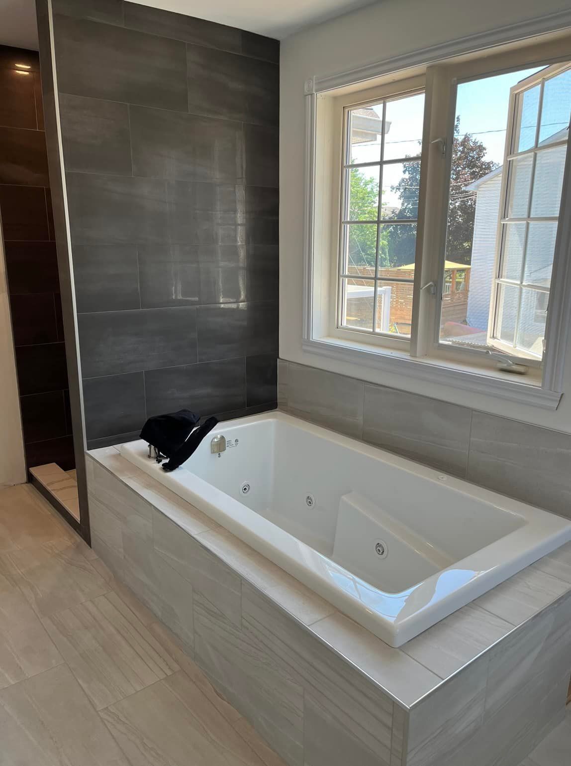 Une salle de bains moderne avec une baignoire balnéo blanche entourée de carreaux gris et une fenêtre donnant sur une journée ensoleillée.