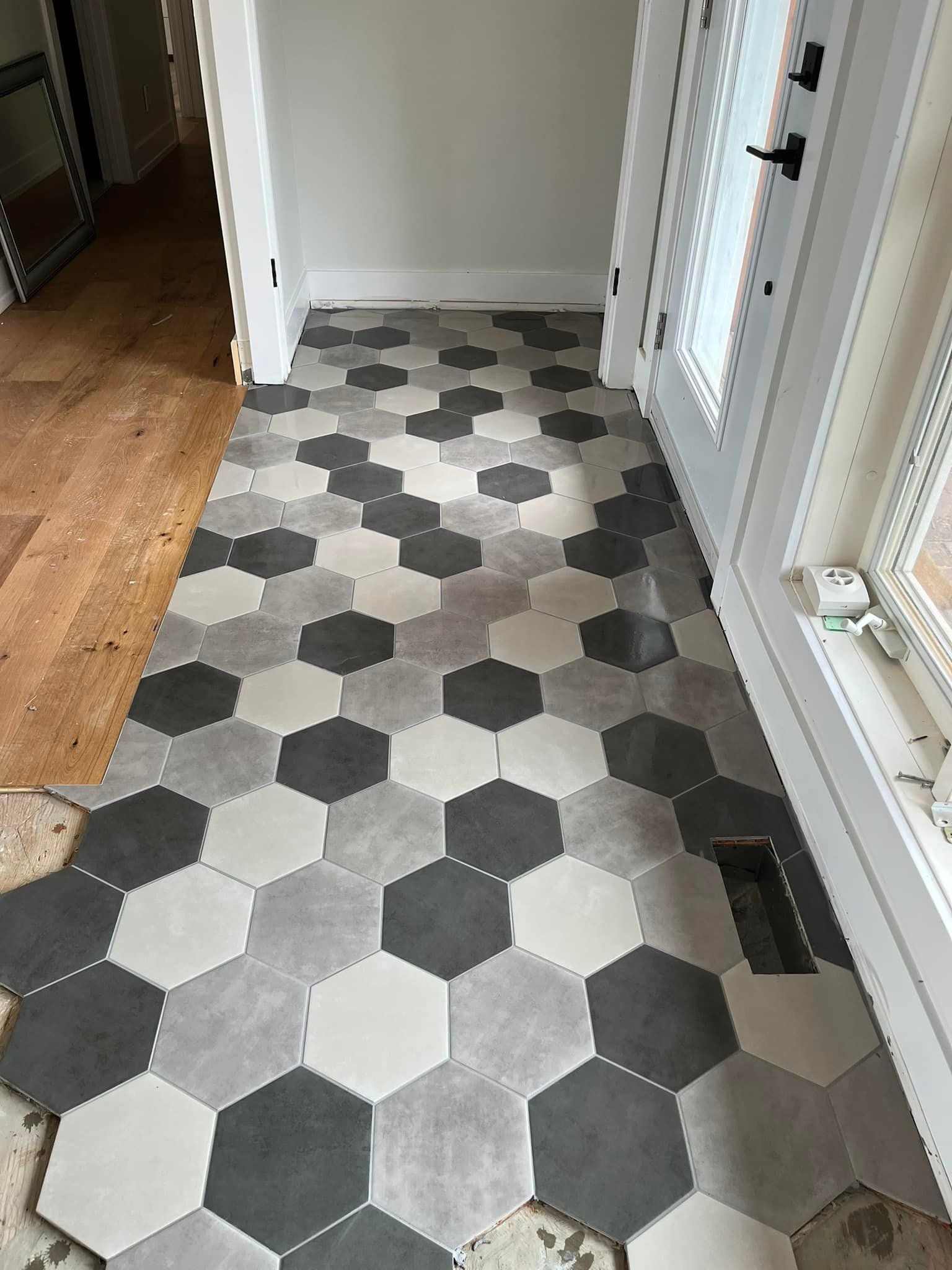 Un sol d'entrée orné d'un motif de carreaux hexagonaux dans des tons de blanc, de gris et d'anthracite, jouxtant un parquet.