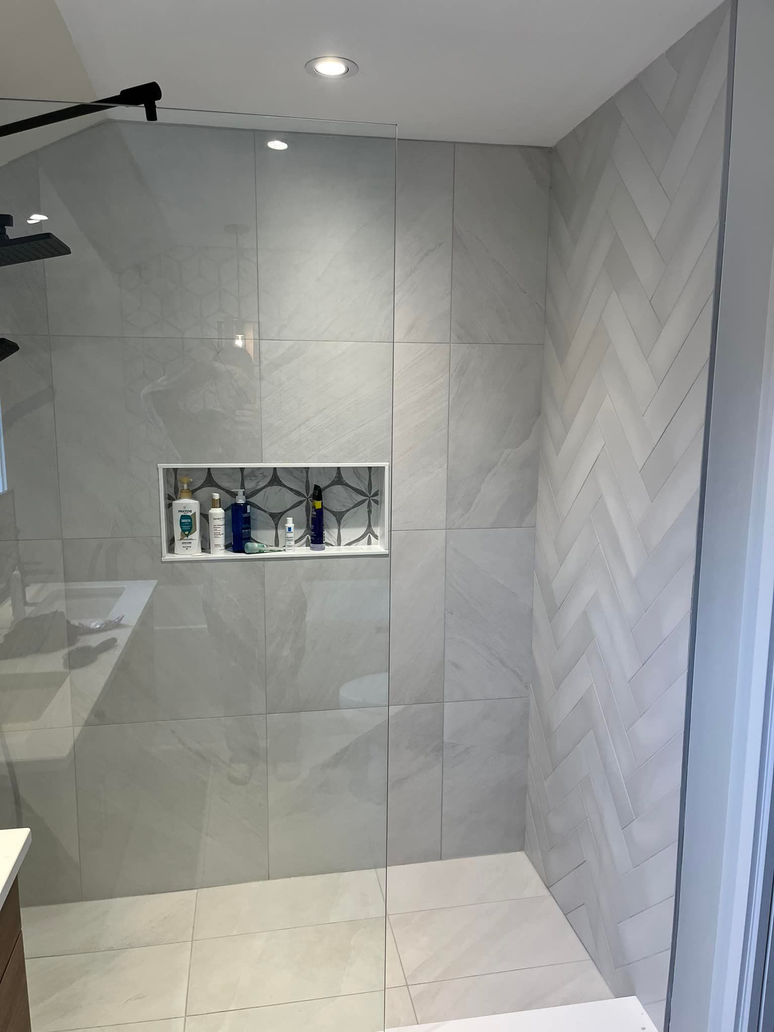 Douche à l'italienne moderne avec grands carreaux gris clair, un mur d'accent en chevrons et une étagère encastrée intégrée avec articles de toilette.