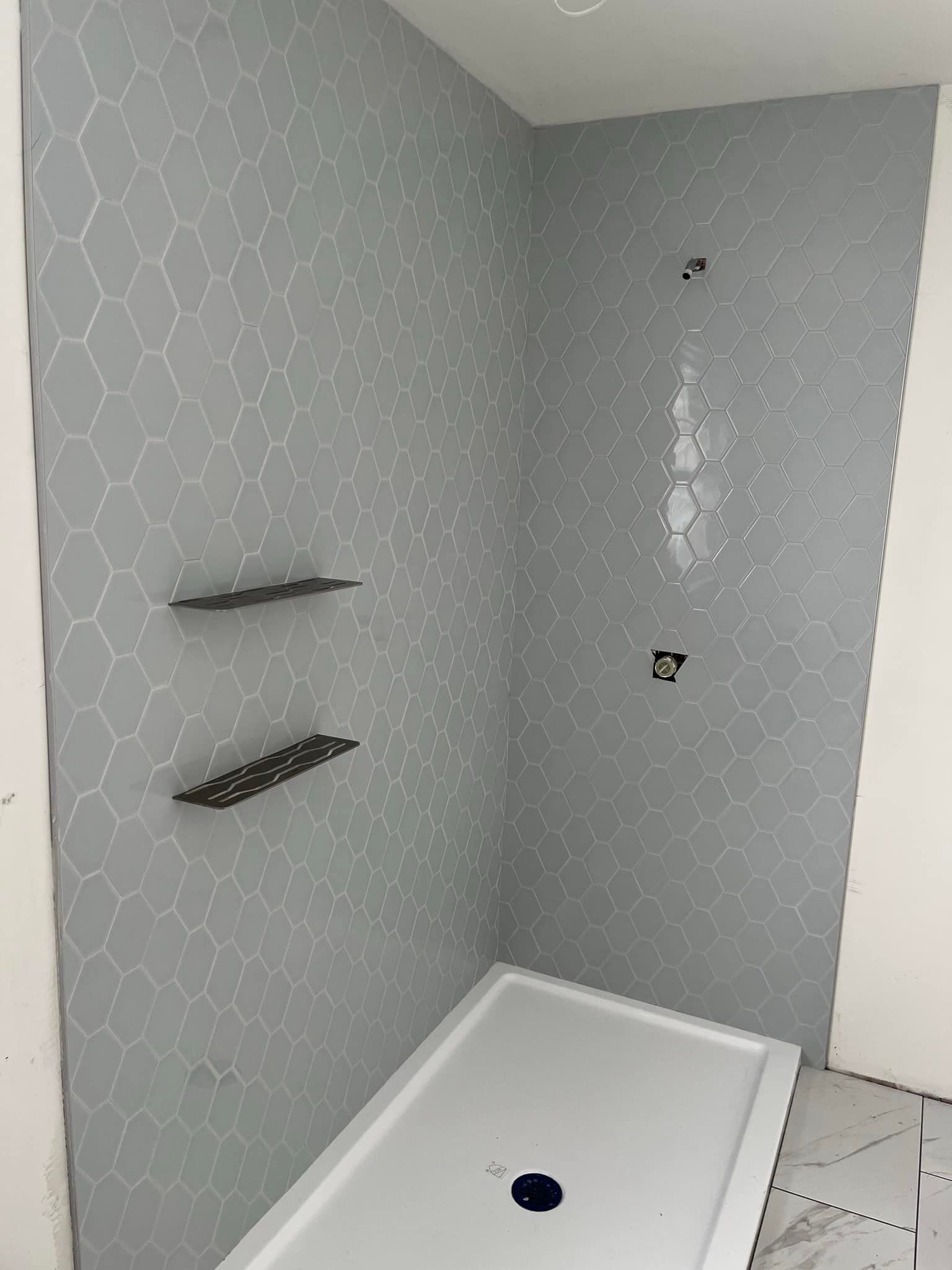 Une cabine de douche avec des carreaux muraux gris à motifs hexagonaux, deux étagères murales et un receveur de douche blanc.