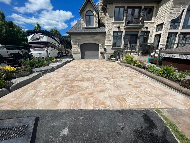 Une allée pavée de pierres beiges mène à une maison en pierre avec une porte de garage cintrée, à côté d'un grand camping-car stationné.