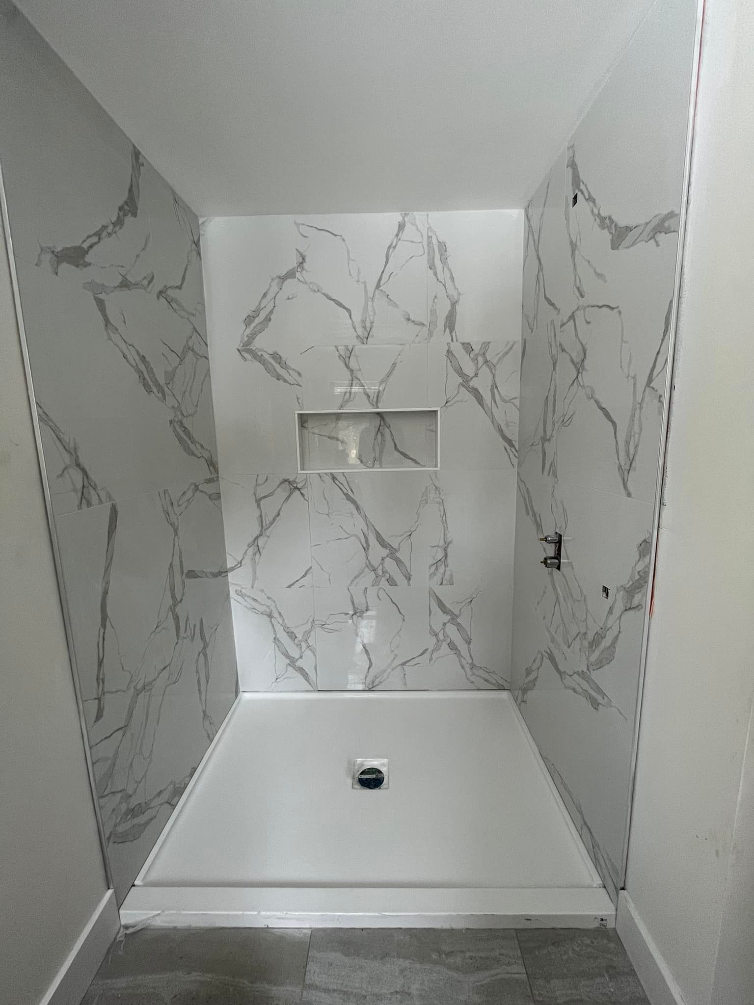 Une douche à l'italienne avec des panneaux muraux en marbre blanc, une étagère centrale encastrée et un receveur de douche blanc.