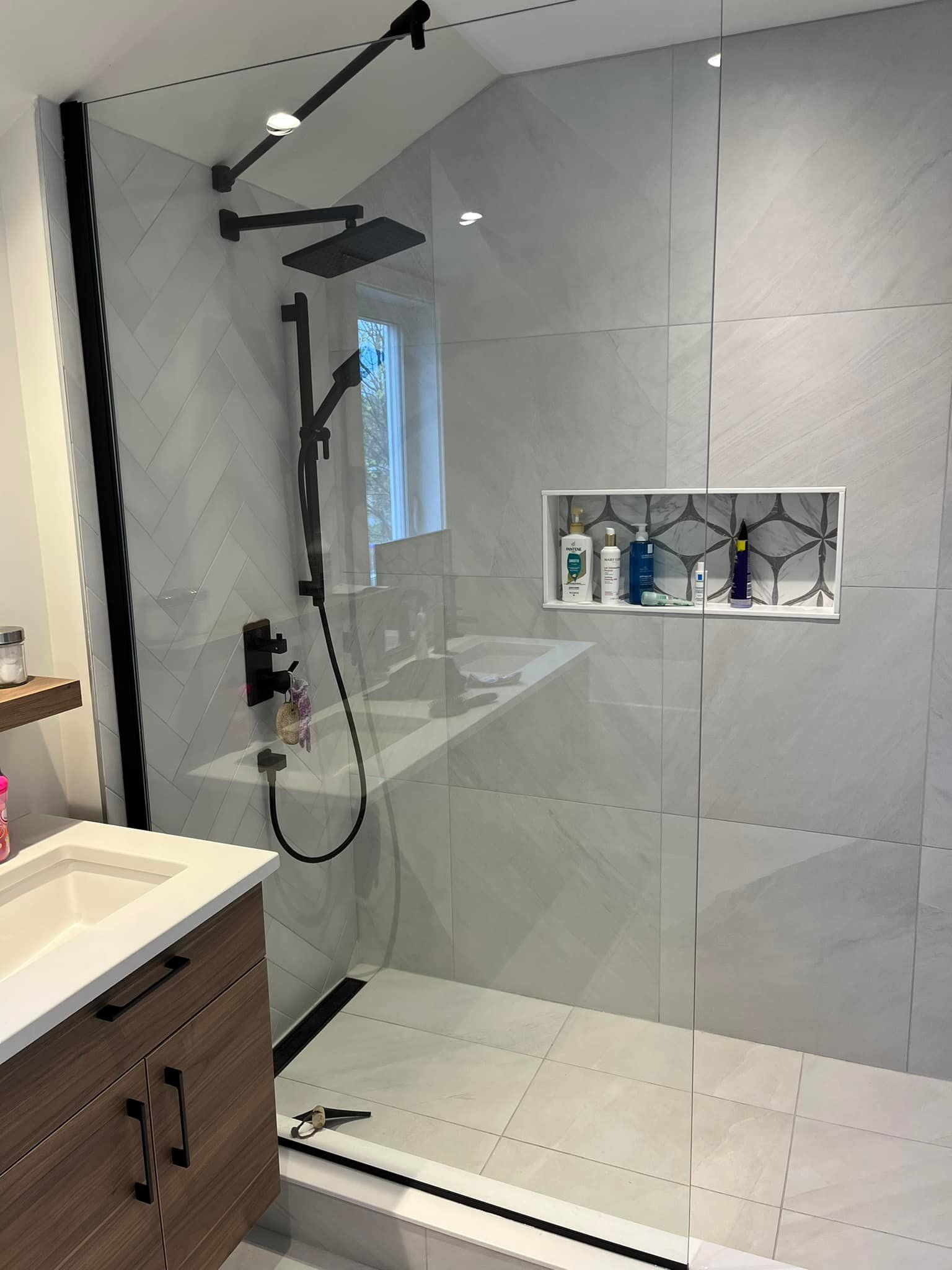 Une douche à l'italienne moderne avec de grands carreaux gris, une pomme de douche effet pluie noire et une niche encastrée avec des articles de toilette.