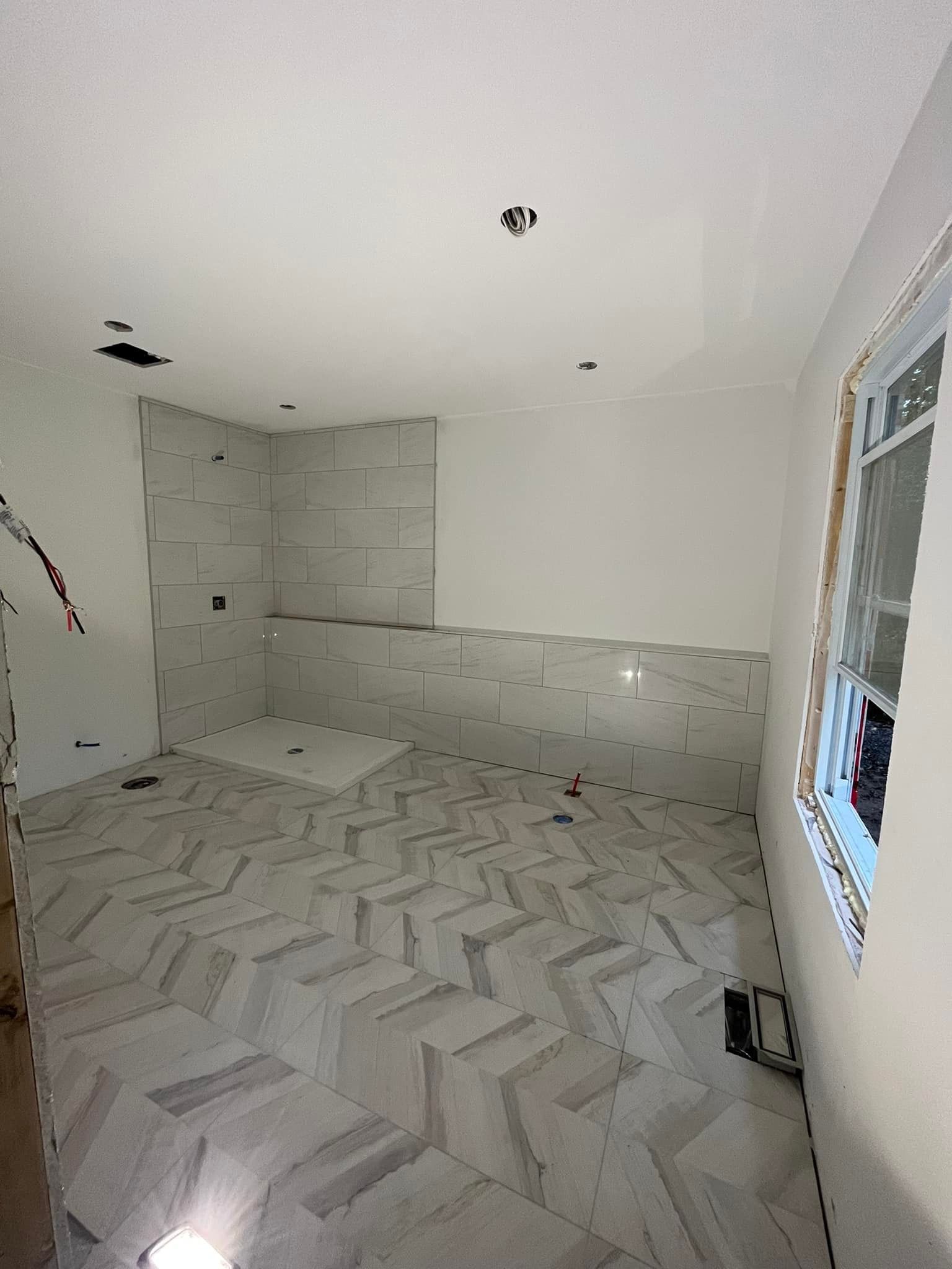 Une salle de bains en construction avec un carrelage au sol à chevrons, des murs de douche partiellement carrelés et une fenêtre.