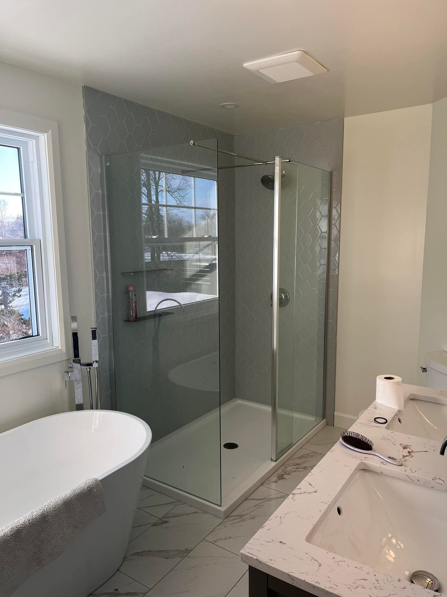 Une salle de bains moderne comprenant une douche à l'italienne en verre, une baignoire blanche sur pieds et une double vasque avec des comptoirs mouchetés.