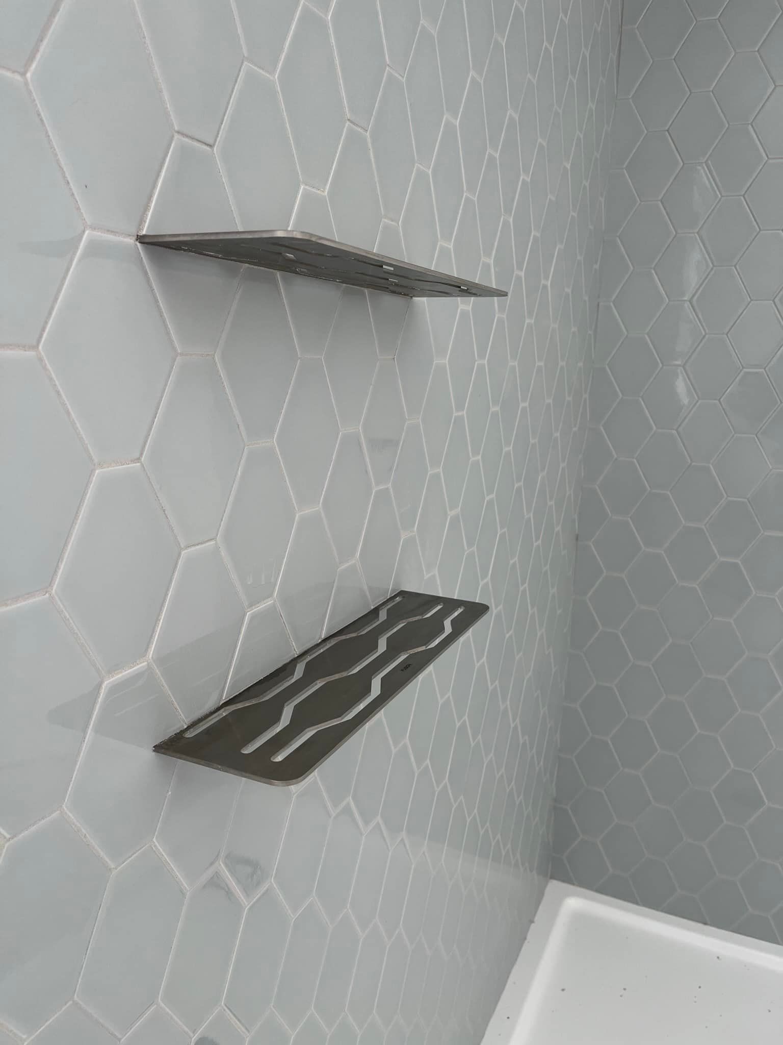 Deux étagères de douche rectangulaires foncées à motif ondulé, fixées sur un mur carrelé hexagonal gris clair.
