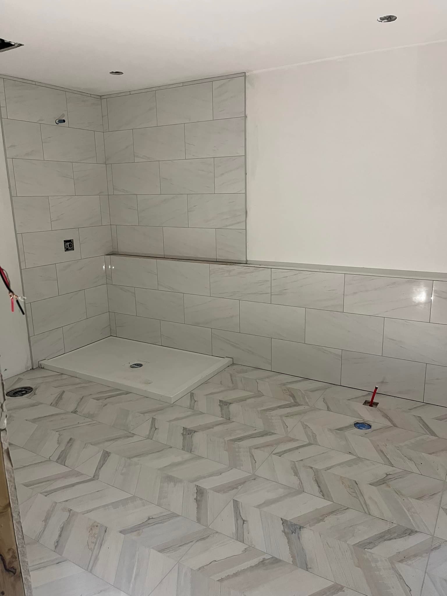 Une salle de bains inachevée avec un carrelage au sol en chevrons et des murs de douche partiellement carrelés de couleur claire.