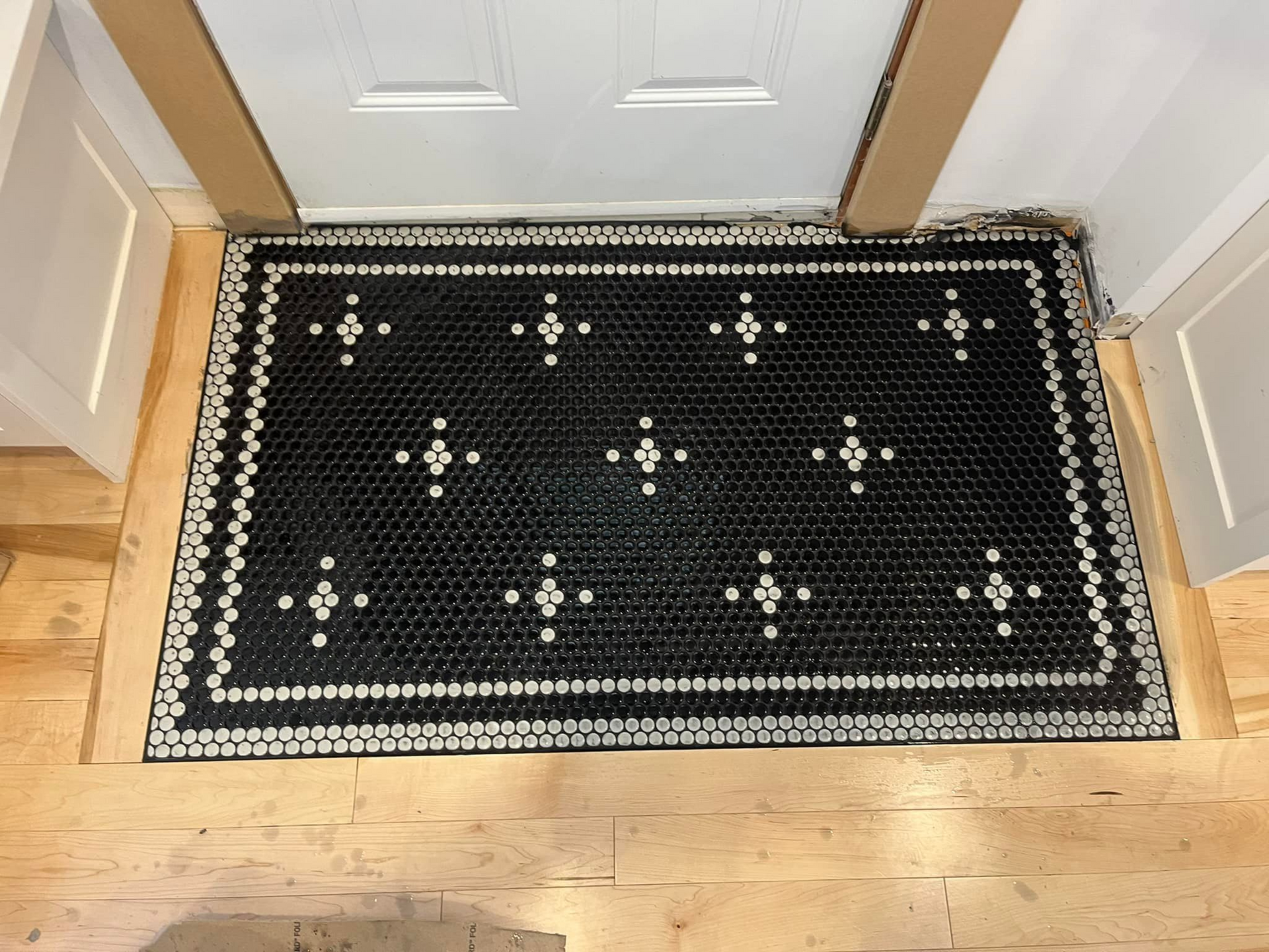 Un tapis rectangulaire noir à bordure de pois blancs et à motifs de croix blanches répétitifs, posé sur un parquet près d'une porte.