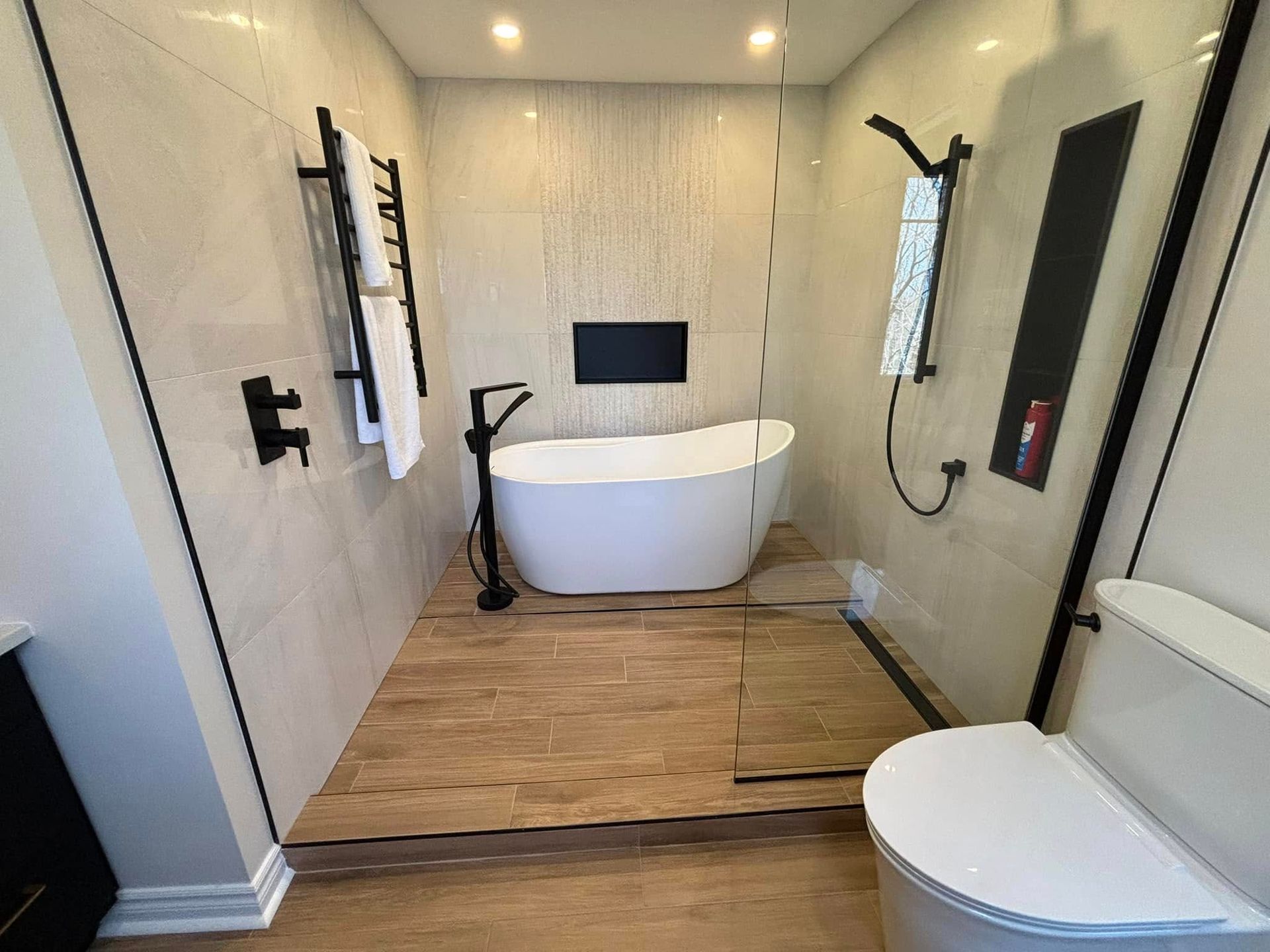 Salle de bains moderne avec baignoire blanche sur pieds sur une plateforme en bois, cabine de douche vitrée et robinetterie noire mate.